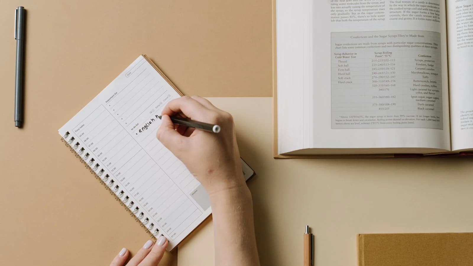 Hands writing in a weekly planner beside an open reference book on a warm neutral desk